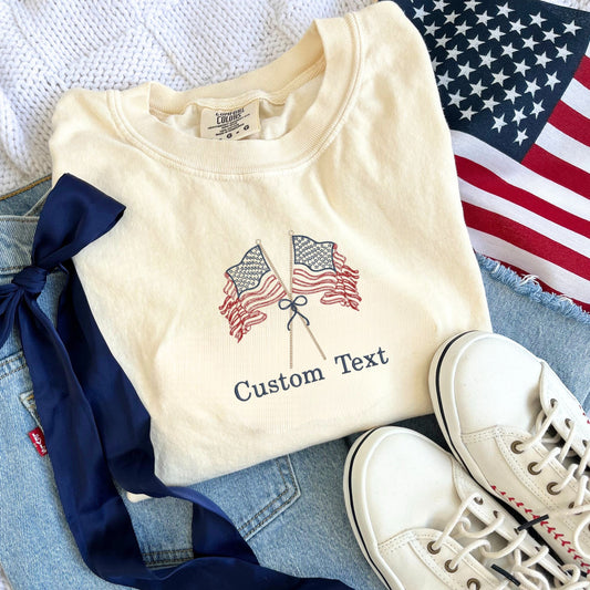 outfit flat lay featuring blue jeans, navy blue ribbon bow, white sneakers, an American flag, and an ivory comfort colors t-shirt with an embroidered crossed stitched American flags on sticks tied with a bow design and custom text underneath