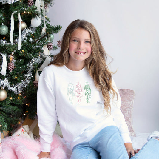young girl wearing a white crewneck sweatshirt with blue, pink, and green embroidered stitched nutcrackers across the chest