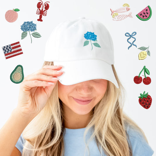 Woman standing in front of a white brick wall wearing a baby blue t-shirt and a white hat with an embroidered hydrangea icon. Girl is surrounded by mockups of other design options such as: lobster, peach, hydrangea, usa flag, avocado, strawberry, cherries, lemon, ribbon bow, watermelon, and sardine.