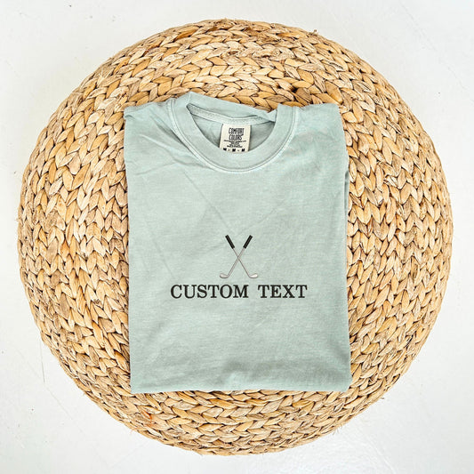 flat lay of bay comfort colors t-shirt with custom text and crossed golf club embroidery on the center chest