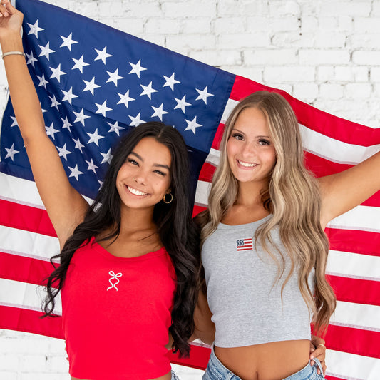 Two women wearing custom embroidered micro spaghetti strap cropped tanks, one in red with a white ribbon bow embroidery and one in gray with a USA flag embroidery, standing in front of an American flag. 4th of July, patriotic, summer fashion, bridal party outfit.