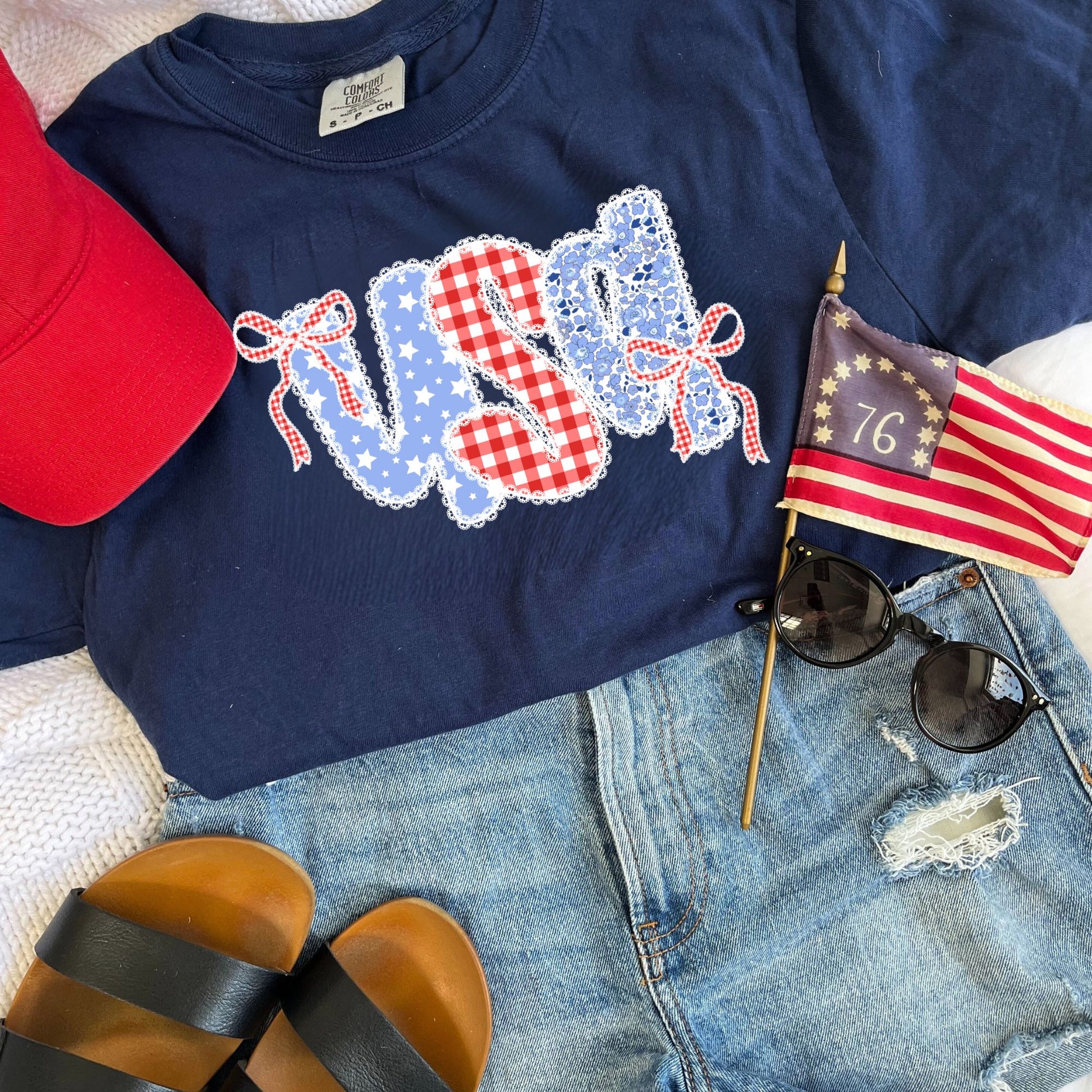 outfit lay out featuring the following: red baseball hat, black and brown Reef sandals, ripped jean shorts, sunglasses, mini 1776 USA flag, and a navy comfort colors t-shirt with coquette bow lace usa floral and gingham print on the center chest
