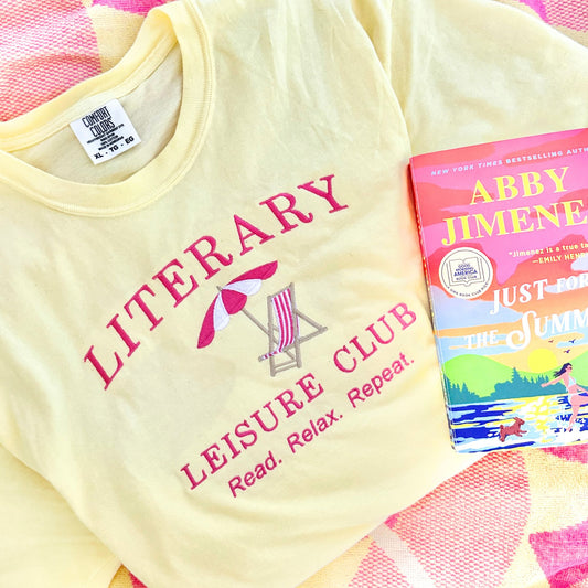 banana comfort colors tee with a beach chair/umbrella and literary leisure club read relax repeat text embroidered design in pink and white thread. T-shirt is folded and laying on a pink and yellow beach towel and Abby Jimenez Just for the Summer book is laying on top of the shirt.