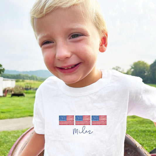 little boy sitting on tractor wearing a white comfort colors t-shirt with custom print featuring 3 traditional american flags with a watercolor look and a name in a script font and blue ink underneath
