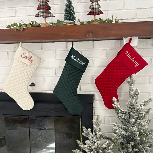 three quilted stockings hanging on a mantle in cream, dark green, and red. On the stockings are embroidered name in different fonts and christmas thread colors.