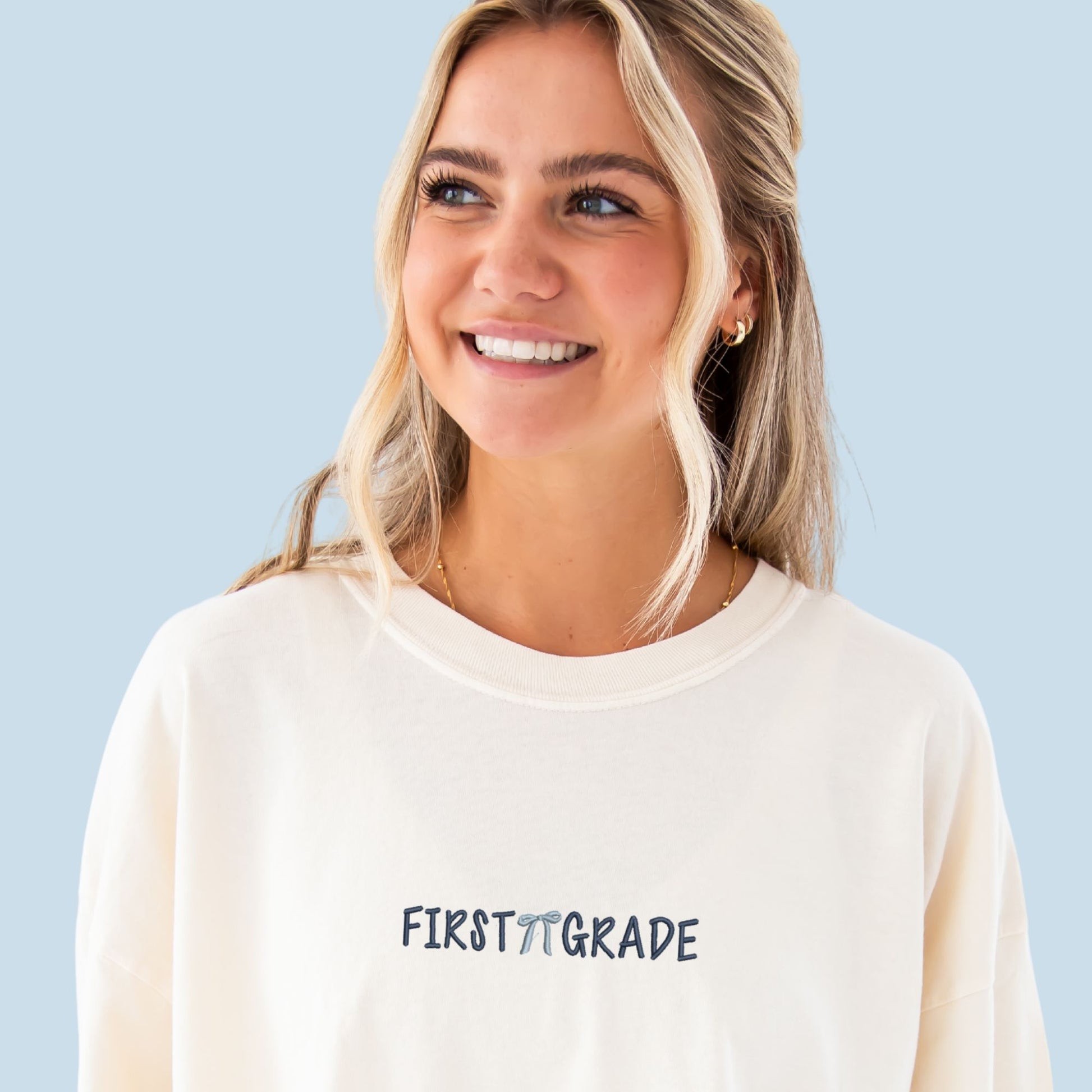 girl standing in front of a blue backdrop wearing an oversized ivory comfort colors t-shirt with embroidered first grade design on the center chest. there's a mini cute bow between first and grade. embroidery is done in navy and powder blue thread