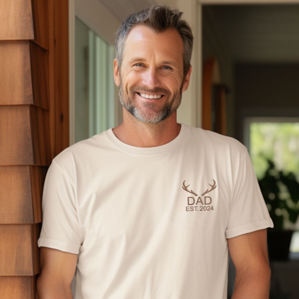 man wearing an ivory t-shirt with custom antler dad date embroidered design on the left chest in chestnut thread