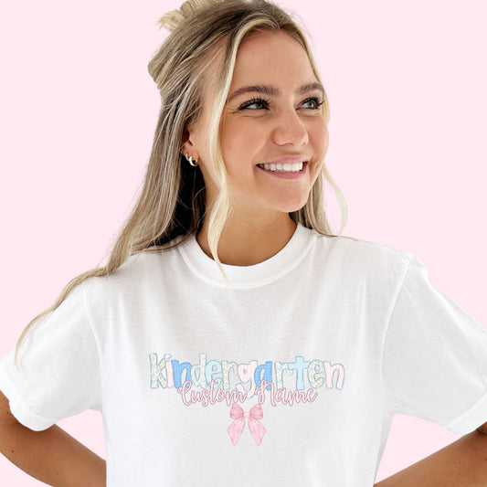 Woman wearing a white t-shirt with 'Kindergarten Custom Name' text and a pink bow graphic on a pink background. Kindergarten is printed with letters that have various background patterns such as  a pastel floral, stripe, and gingham.
