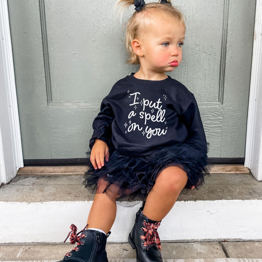 little girl wearing cute boots and a tulle skirt and a black crewneck sweatshirt  with a cute I put a spell on you wavy script printed design