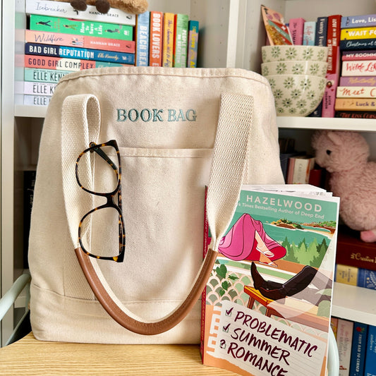 Canvas tote bag with 'BOOK BAG' embroidered in eucalyptus thread on the front above the pocket, resting on a wooden surface. A pair of tortoiseshell reading glasses hangs from the front pocket, handles have a brown faux leather accent. A paperback book titled Problematic Summer Romance by Hazelwood leaned against the front. In the background are bookshelves filled with colorful romance novels, a stack of floral bowls, and a pink teddy bear