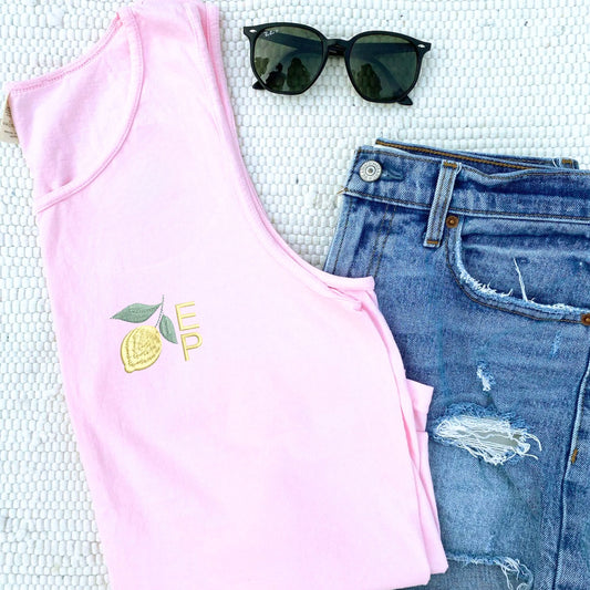 Flat lay of folded blossom tank with lemon icon embroidered on the left chest with custom monogram beside the icon. partnered with denim shorts and sunglasses.