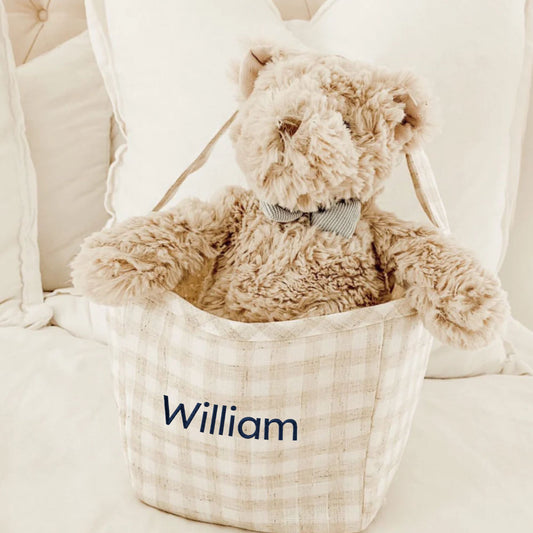 Teddy bear in a basket with 'William' embroidered on it, sitting on a white surface.
