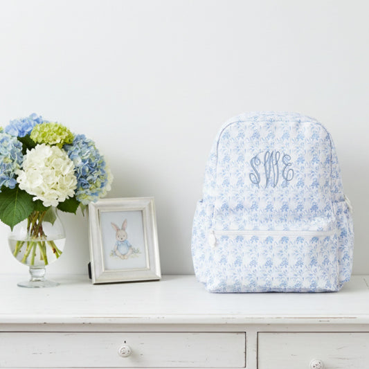 Navy floral-patterned backpack with monogram on a white surface with flowers and a picture frame.