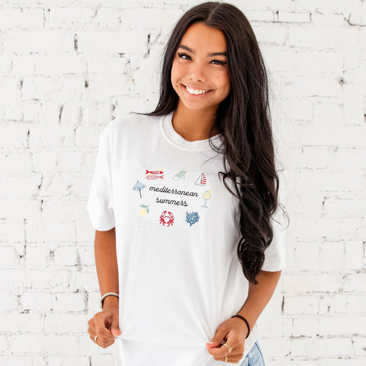 girl standing in front of a white brick wall with an embroidered design on the front center featuring: sardines, an umbrella, beach chair, sailboat, cocktail, coral, crab, and a lemon icon all surrounding Mediterranean summers text in black thread