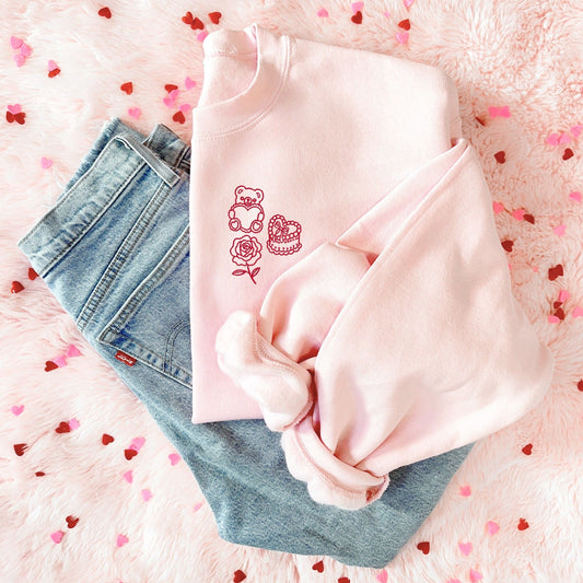 Pink sweatshirt with a coquette valentines icons (teddy bear, heart cake, rose) design embroidered on the left chest in red thread, paired with jeans against a pink background with heart shapes