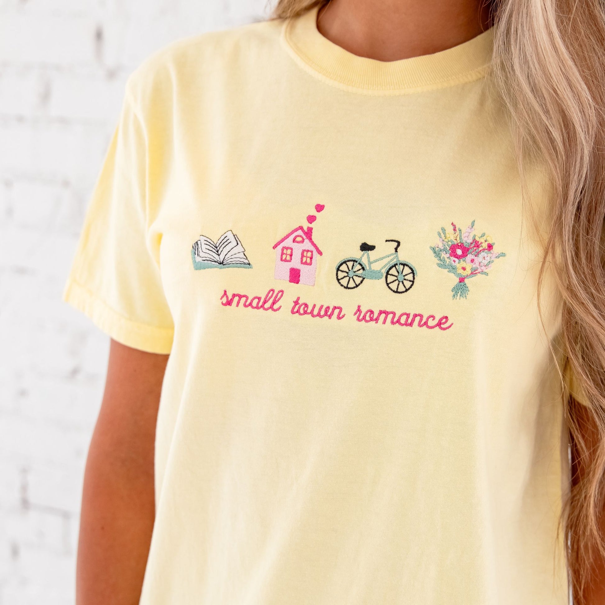 close up of a girl with long blonde hair wearing a banana comfort colors t-shirt with a custom small town romance design embroidered on the center front featuring an open book, house with hearts coming out of the chimney, bike, and flower bouquet icons.