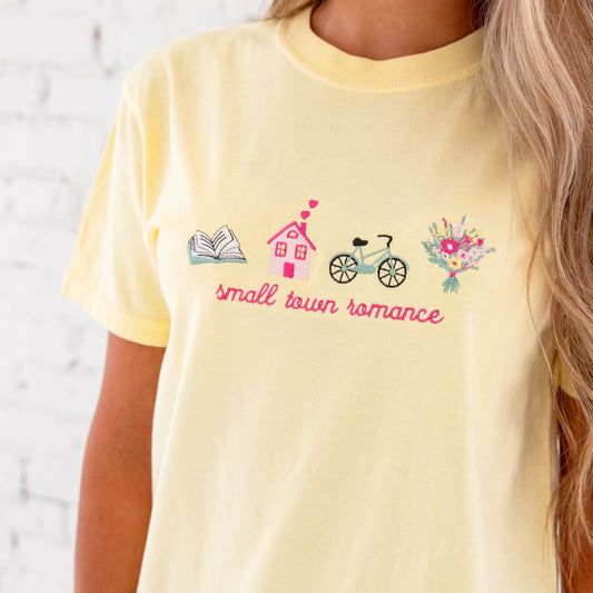 close up of a girl with long blonde hair wearing a banana comfort colors t-shirt with a custom small town romance design embroidered on the center front featuring an open book, house with hearts coming out of the chimney, bike, and flower bouquet icons.