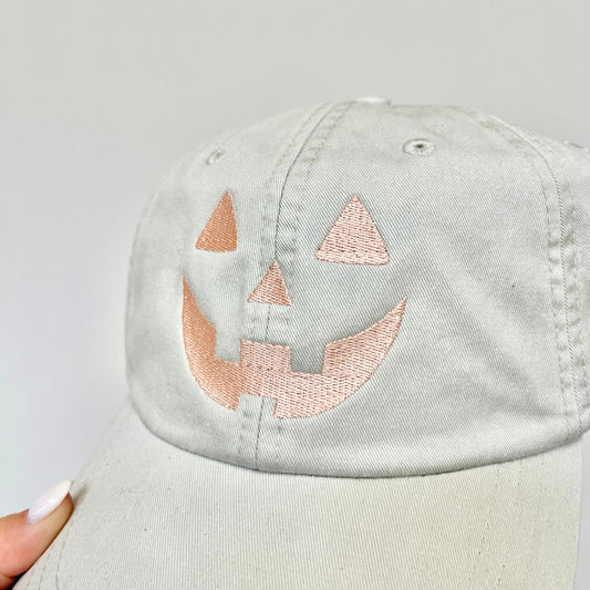Ivory baseball cap with a pumpkin face embroidered design held up in front of a  light gray wall
