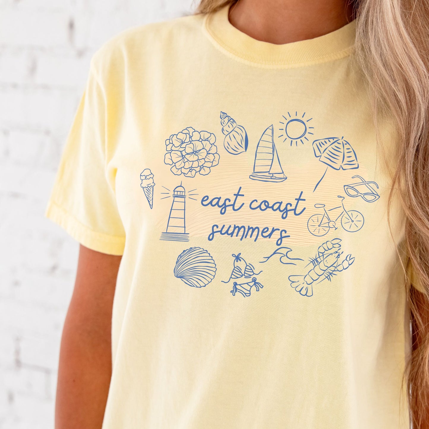 Close up of a woman wearing a banana comfort colors shirt with a custom design featuring a flower, shell, sailboat, sun, umbrella, lighthouse, clam, swimsuit, waves, lobster, bicycle, sunglasses, and east coast summer printed at the center in a script font.