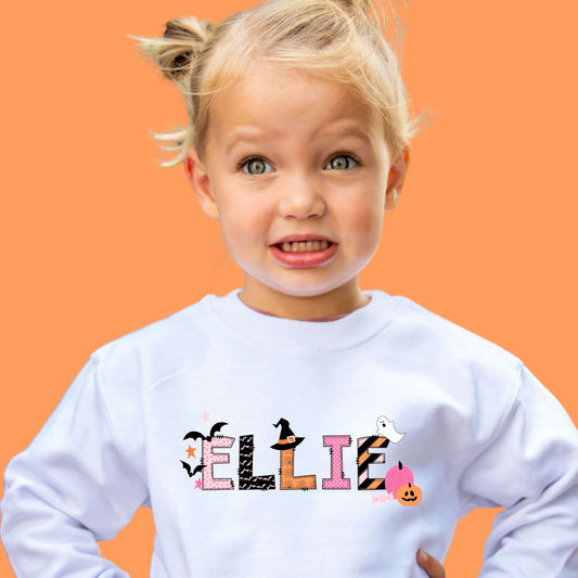 Little girl wearing a white crewneck sweatshirt with a custom name print in mix of pink, orange, and black halloween patterned text decorated with cute spooky season icons.
