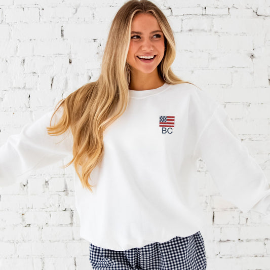 Woman standing in front of brick wall wearing sweet cream sweatshirt with USA Flag icon embroidered at the left chest with custom monogram design embroidered below the USA Flag. Partnered with blue and white gingham shorts.