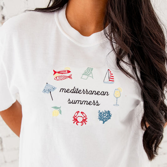girl standing in front of a white brick wall with an embroidered design on the front center featuring: sardines, an umbrella, beach chair, sailboat, cocktail, coral, crab, and a lemon icon all surrounding Mediterranean summers text in black thread
