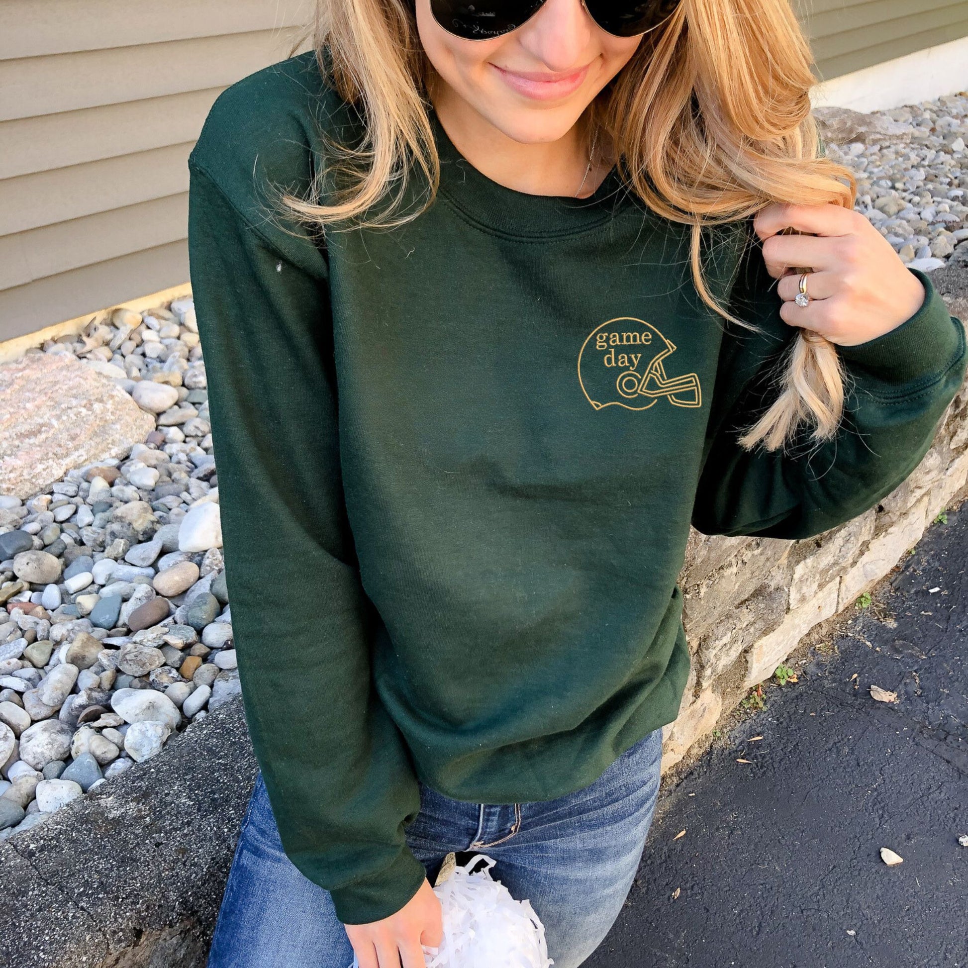 woman wearing a forest green crewneck with embroidered game day and football helmet on the left chest in gold thread