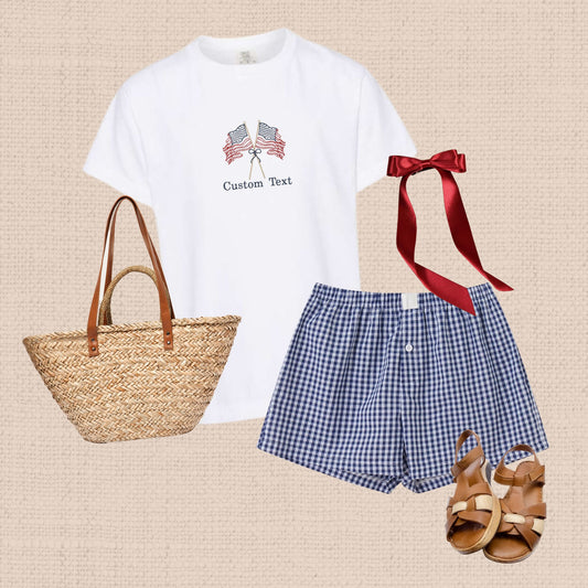 outfit flat lay featuring blue gingham boxer shorts, red ribbon bow, brown sandals, straw tote bag, and a white comfort colors t-shirt with an embroidered crossed stitched American flags on sticks tied with a bow design and custom text underneath
