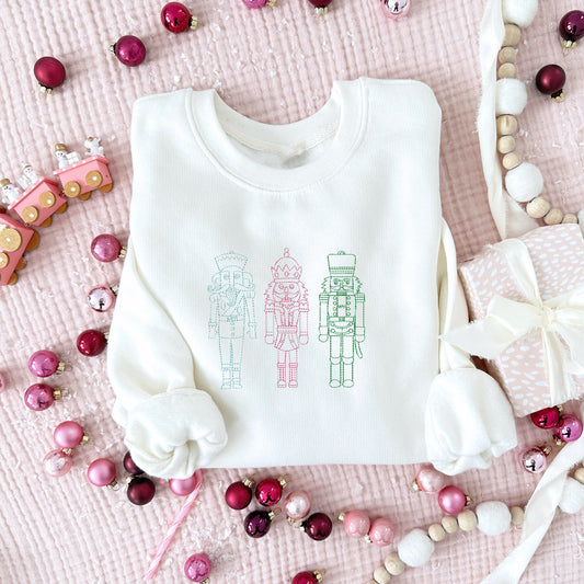 pink and fun Christmas styled flat ay of a young white crewneck sweatshirt with blue, pink, and green embroidered stitched nutcrackers across the chest