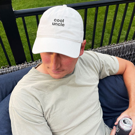 Man sitting on his porch wearing a stone baseball cap with cool uncle embroidered in a navy thread