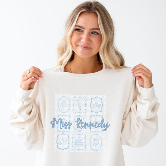 woman wearing a sweet cream crewneck sweatshirt with custom print. print features a pale blue gingham pattern background with nine bow frames with cute line sketch teacher icons. Also features custom script text name across the center of the design in a darker blue ink.