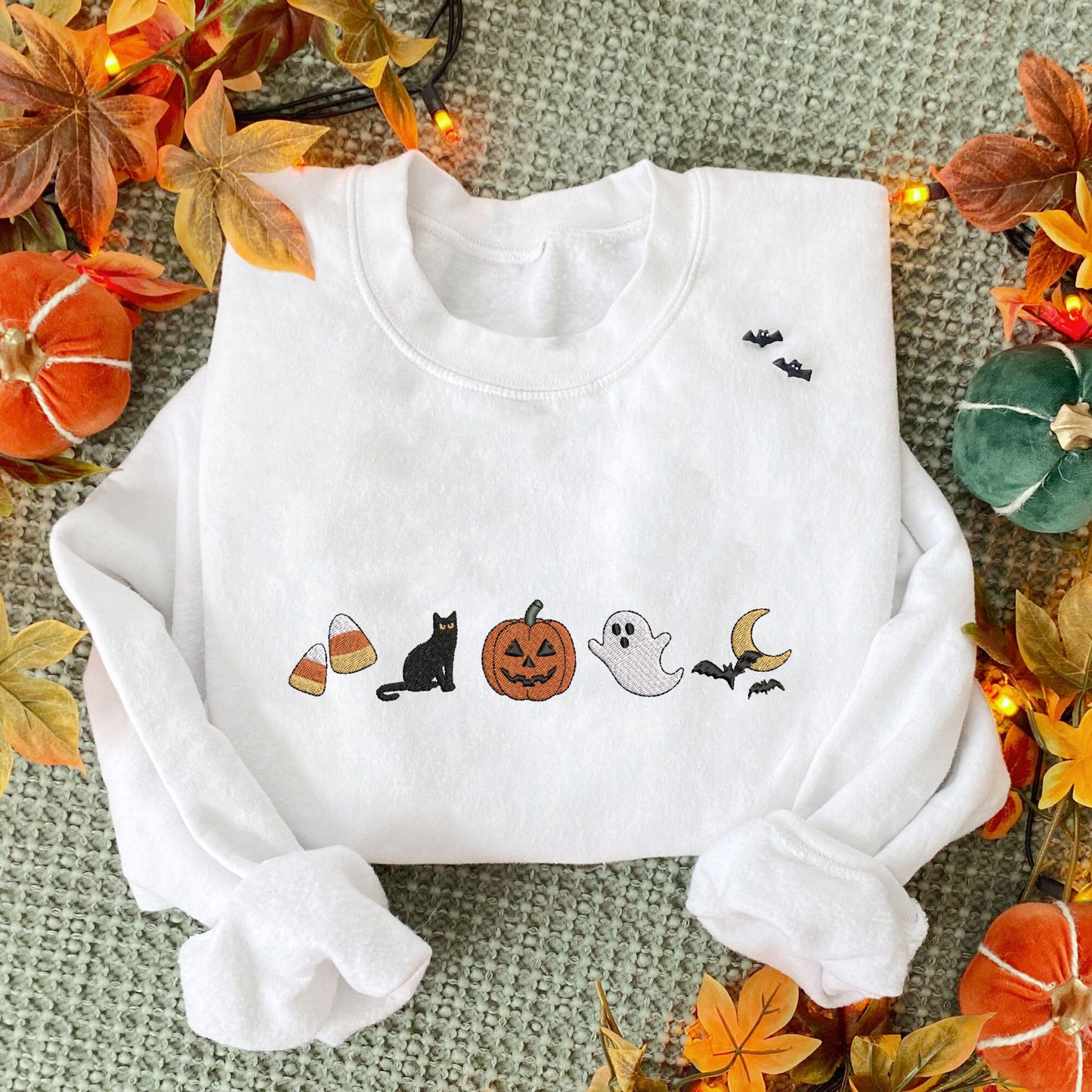 styled fall flat lay photo of a white crewneck sweatshirt with embroidered halloween icons. The icons are embroidered as following: candy corn, black cat, jack-o-lantern, ghost, and bats flying over the moon.