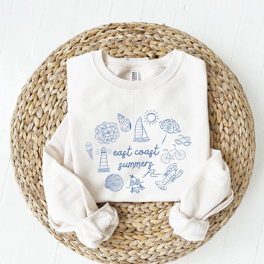 folded sweet cream sweatshirt with flower, seashell, sailboat, sun, umbrella, sunglasses, bicycle, lobster, wave, swimsuit, clam, lighthouse, ice cream and east coast summers script text printed on the middle text.