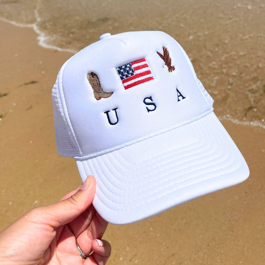 a white trucker hat with custom cowboy boot, usa flag, and bald eagle USA embroidered design held above the sand on the beach