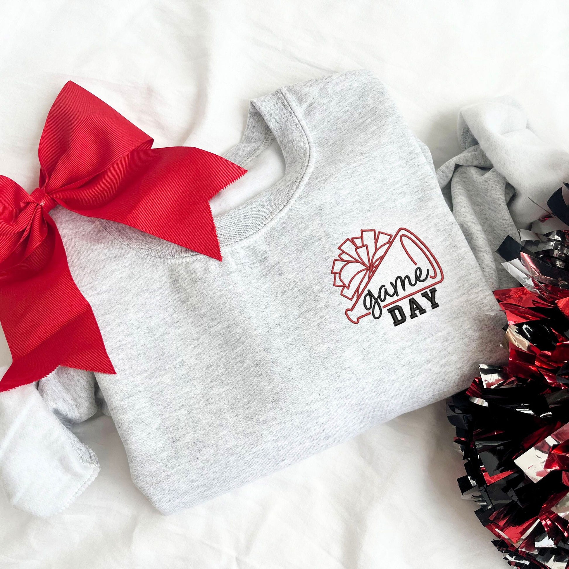 flat lay of an ash crewneck sweatshirt with embroidered cheer megaphone and pom pom with text game day on the left chest in red and black threads.