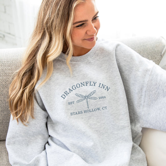 Woman wearing an oversized ash sweatshirt with Dragonfly Inn Stars Hollow, CT embroidered design in French Blue thread