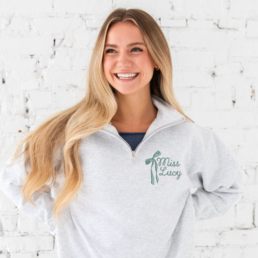 woman standing in front of a white brick wall wearing an ash quarter zip sweatshirt with a custom embroidered design on the left chest featuring a a long tail bow next to a curly script name in eucalyptus green thread
