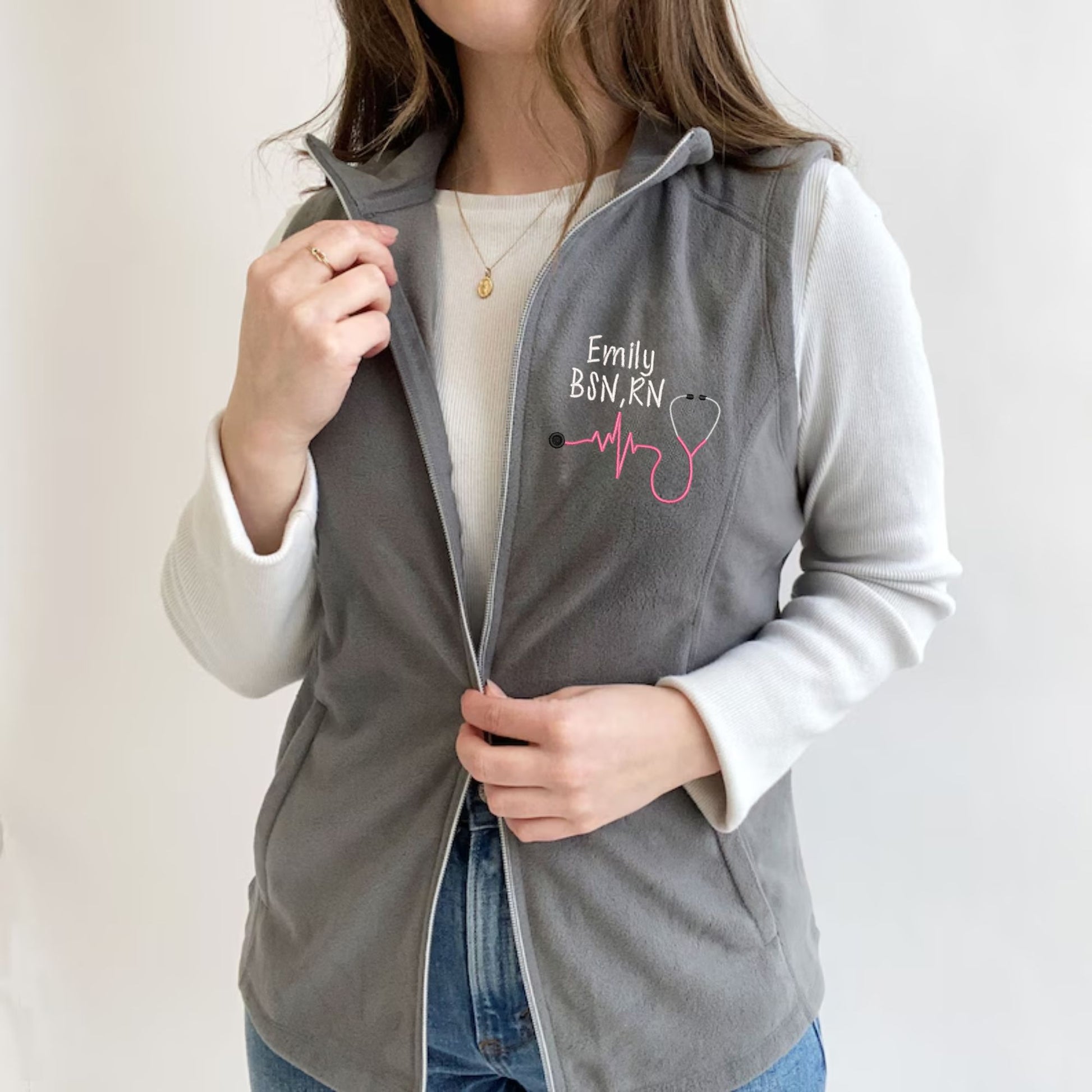 young woman wearing a light weight grey fleece vest with embroidered nurse heartbeat stethoscope in pink thread  on the left chest 
 with name and credentials in white thread.