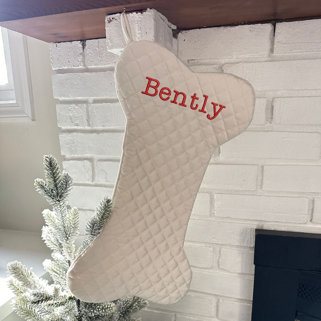 cream quilted Christmas stocking in the shape of a dog bone with embroidered name Bently in red near the top of the stocking