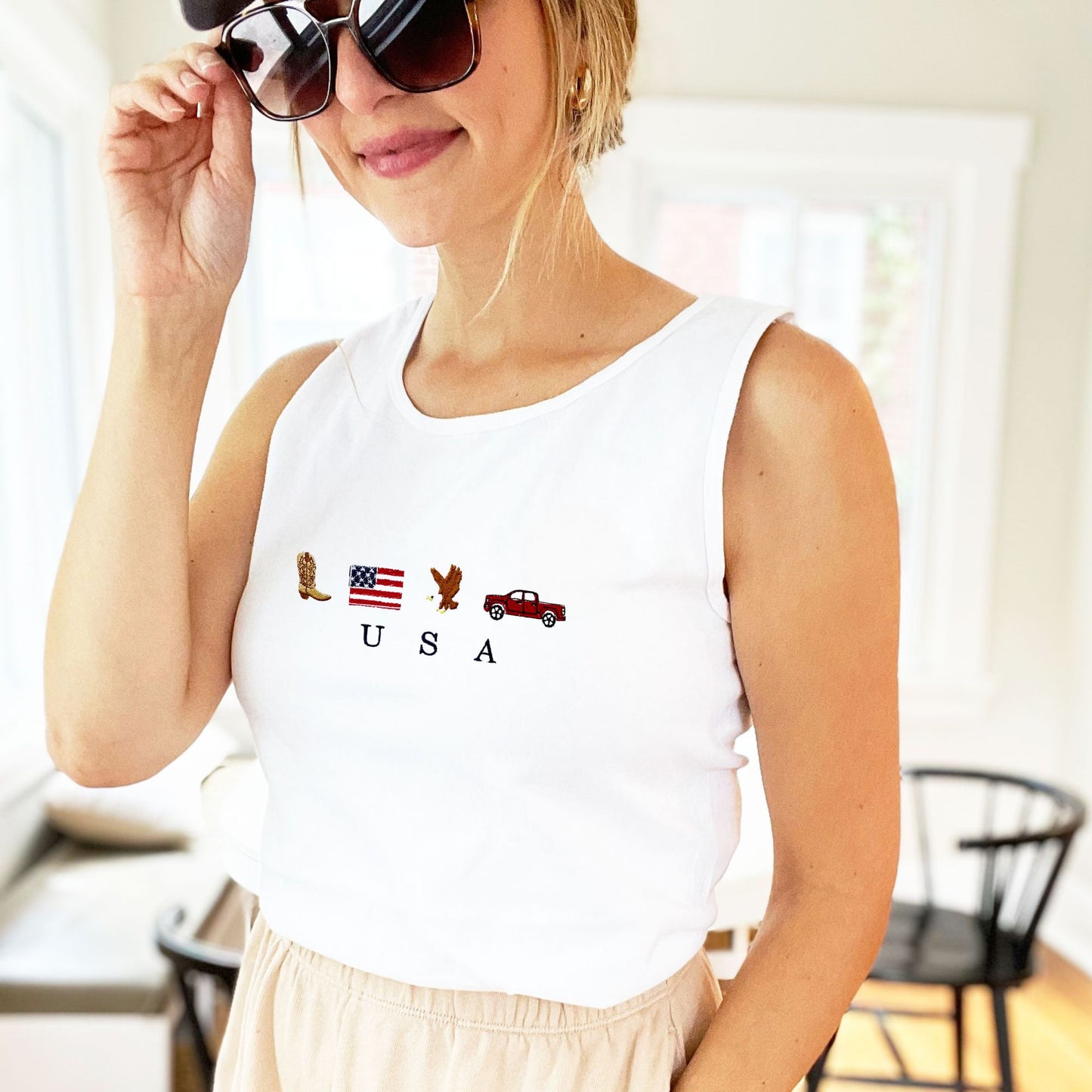 woman wearing a white comfort colors tank top with custom usa icon embroidered design on the center chest featuring a cowboy boot, american flag, bald eagle, and a pickup truck