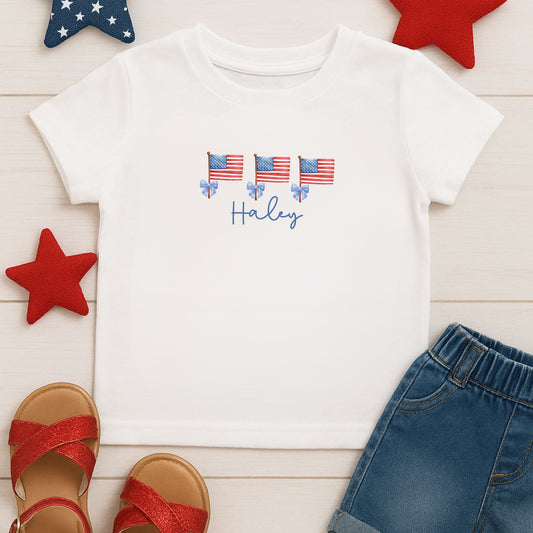 White wood backdrop with blue and red stars, white kids comfort colors t-shirt with a custom print featuring watercolor american flags on a pole with a bow and name in script font underneath paired with red glitter sandals and blue jean shorts.