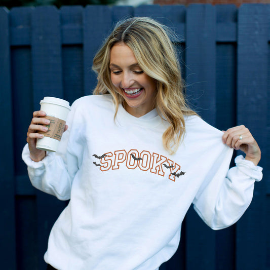 woman wearing a white crewneck sweatshirt with embroidered SPOOKY and bats design in pumpkin spice and black threads