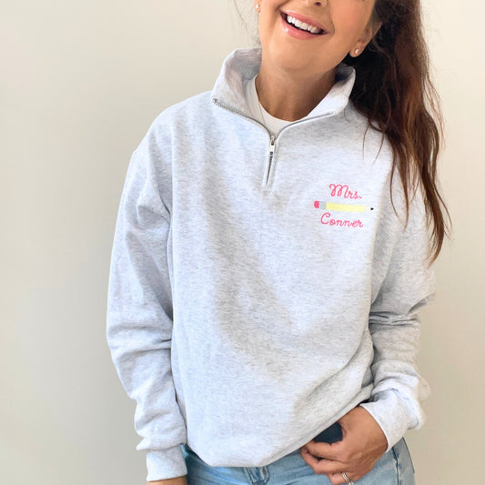 woman wearing an ash quarter zip sweatshirt with a custom curly script name and pencil embroidered design on the left chest