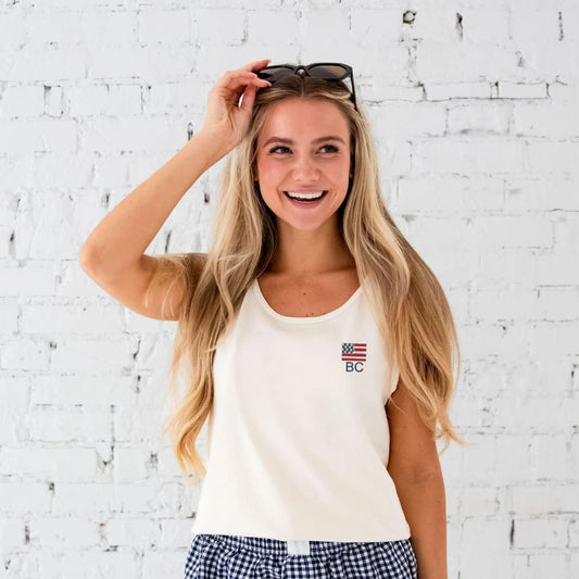 woman standing in front of brick wall, wearing ivory tank with USA flag icon embroidered on the left chest with custom monogram below the icon. partnered with blue and white gingham shorts