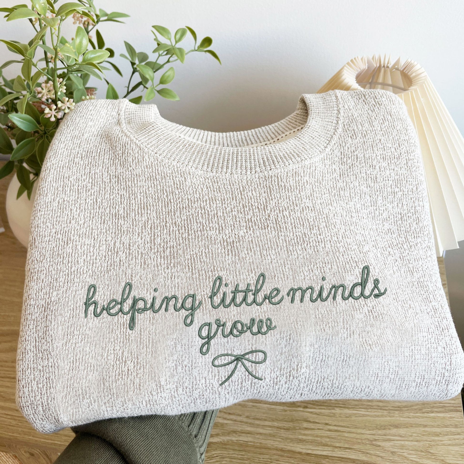 latte cozy knit nantucket fleece sweatshirt folded and held out above a light wooden dresser. The sweatshirt features a cute embroidered helping little minds grow design in a script font with a small delicate bow underneath. Embroidery is in eucalyptus thread.