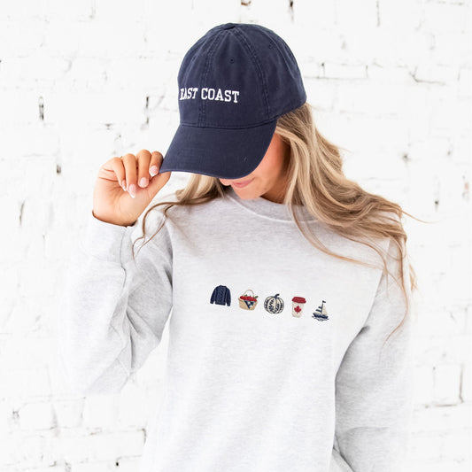 A woman wearing a navy blue baseball cap with 'EAST COAST' embroidered in white and a light heather gray sweatshirt with a small embroidered east coast fall nautical-themed graphic on the chest, posed against a white brick wall.