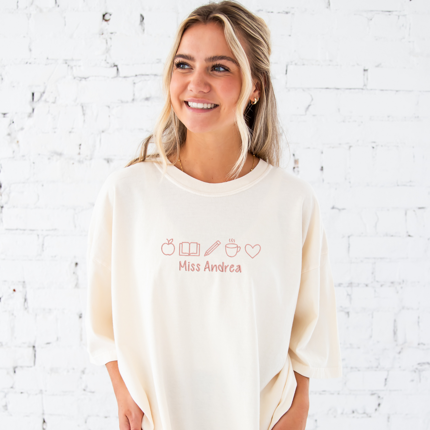 young woman wearing an ivory comfort colors t-shirt with embroidered teacher icons in coral pink thread across the chest and custom text underneith