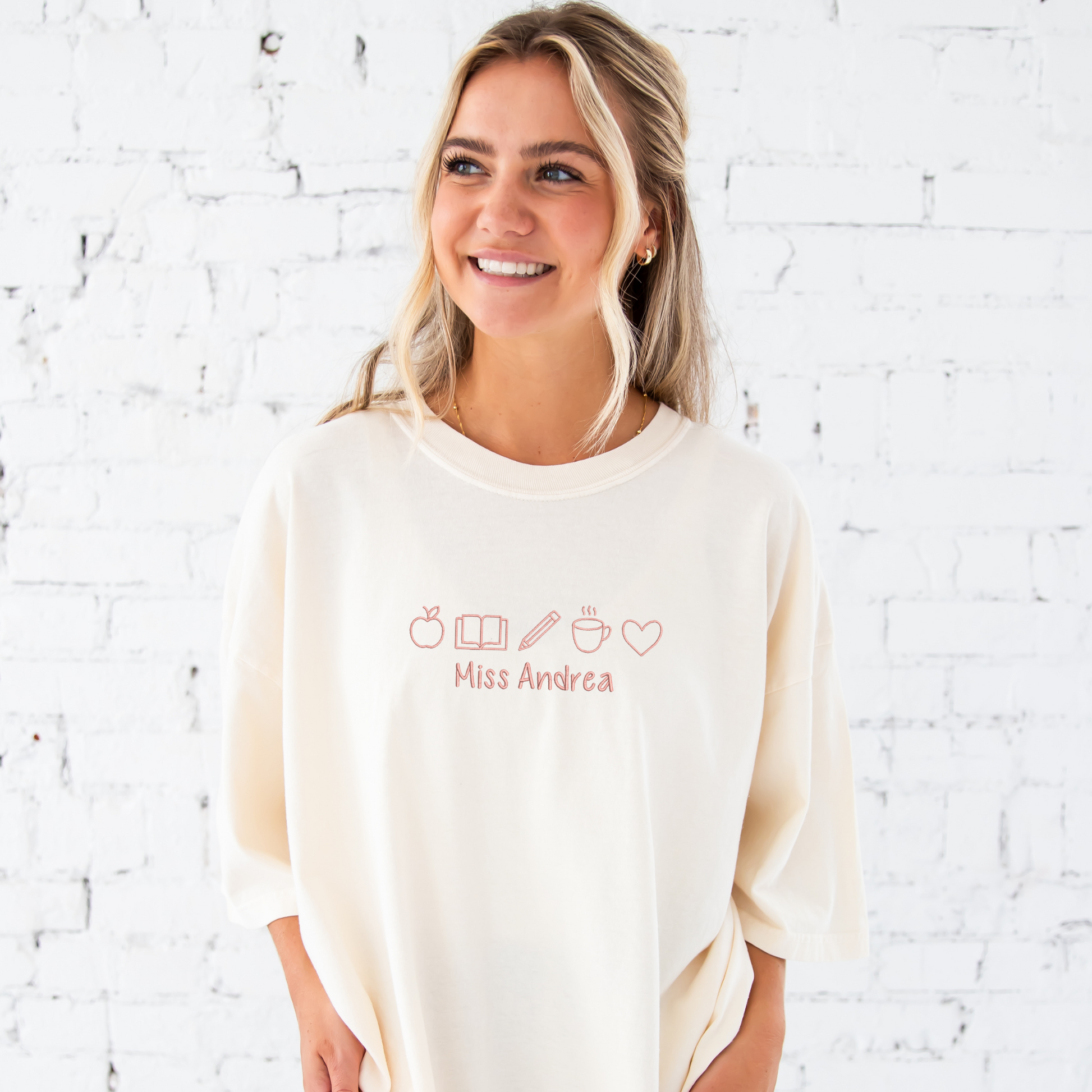 young woman wearing an ivory comfort colors t-shirt with embroidered teacher icons in coral pink thread across the chest and custom text underneith
