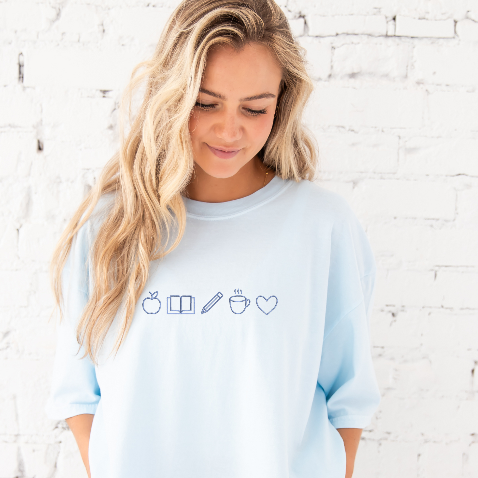 young woman wearing chambray tee with embroidered teacher icons in periwinkle thread across the chest