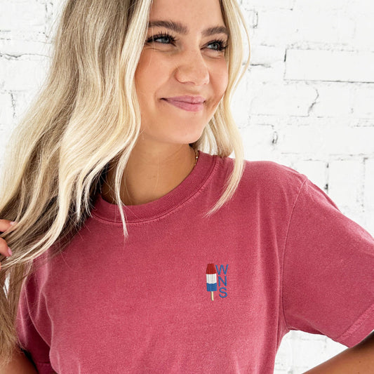 Girl standing in front of a white brick wall wearing a crimson comfort colors t-shirt with a custom embroidered design on the left chest featuring a mini red, white, and blue popsicle icon and 3 initials